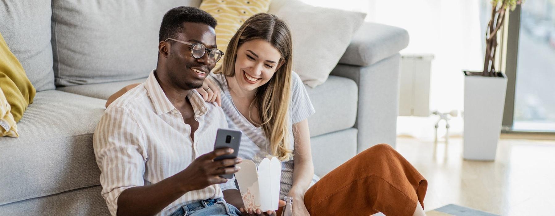 a man and woman with take out, sit against a couch on their living room floor and watch at their cell phone