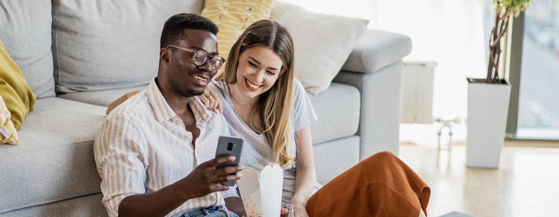 a man and woman with take out, sit against a couch on their living room floor and watch at their cell phone